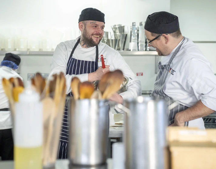 A chef with tattoos and a striped apron smiles while chatting with an instructor in a busy kitchen, with cooking utensils and supplies in the foreground. A chef with tattoos and a striped apron smiles while chatting with an instructor in a busy kitchen, with cooking utensils and supplies in the foreground.